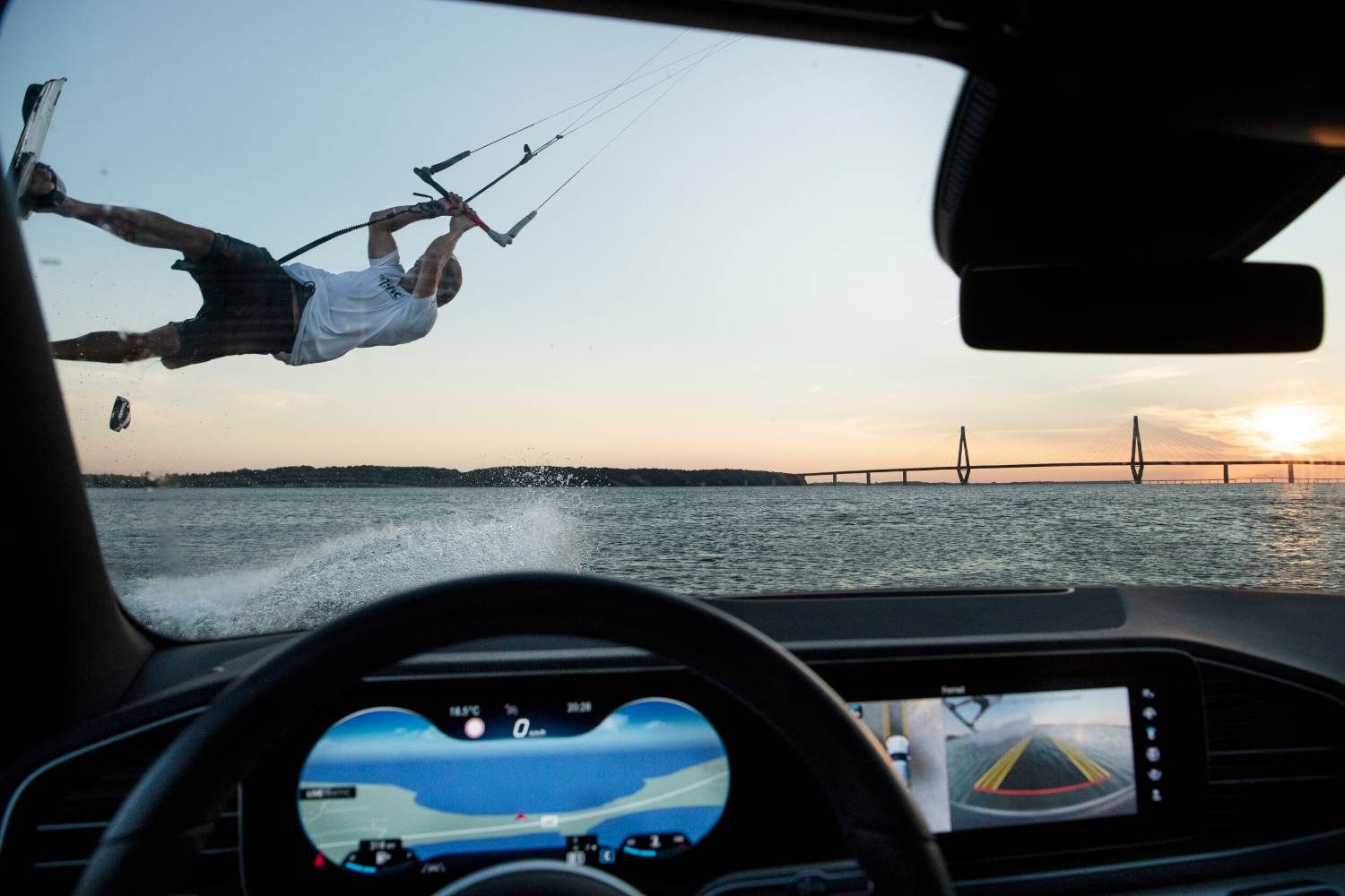 Kitesurfer performing a jump over water, viewed from inside a car at sunset.
