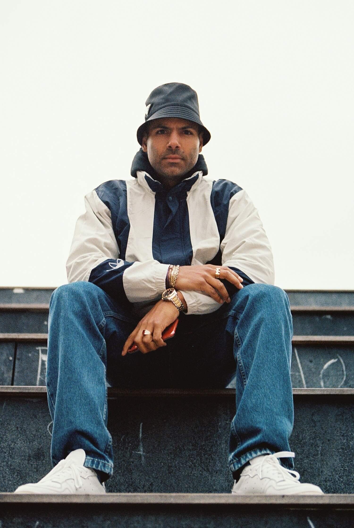 Mads Emil Møller sits on stairs, wearing a bucket hat and casual attire, looking confidently at the camera.