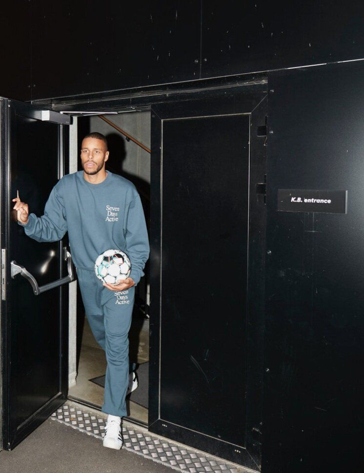 Mathias Zanka exits a dark doorway, holding a soccer ball and gesturing.