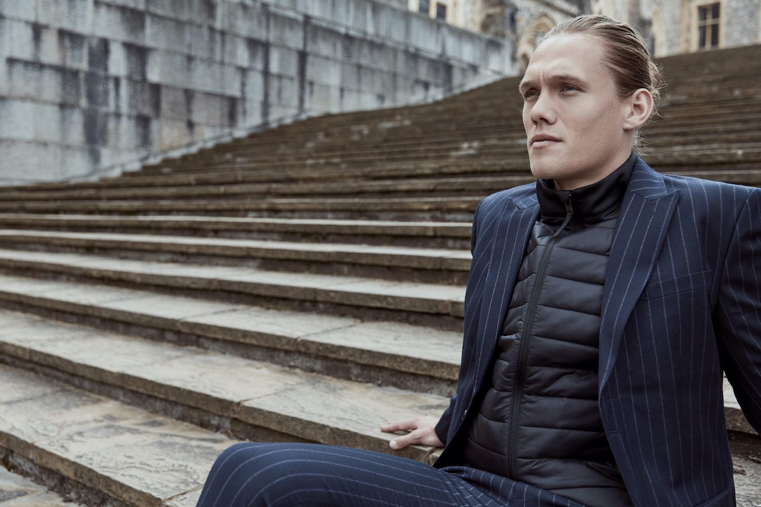 Jannik Vestergaard seated on stone steps, wearing a pinstriped suit and puffer jacket.