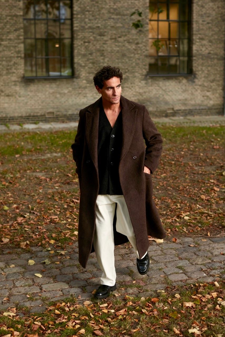Jonathan Harboe walks through a park, wearing a brown coat and white trousers.