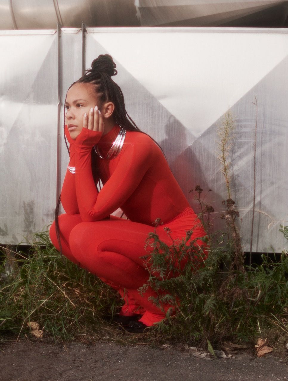 Bathsheba poses thoughtfully in a red outfit, crouched beside greenery and metallic backdrop.