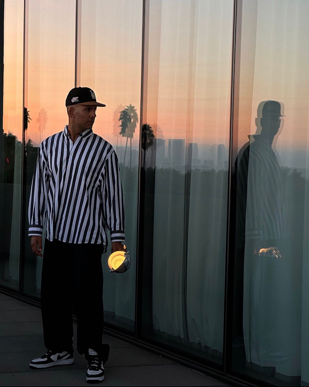 Mads Emil Møller stands by glass reflecting a sunset, wearing a striped shirt and cap.