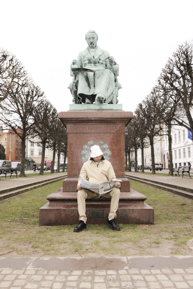 Mads Emil Møller sits reading a newspaper in front of a statue in a park.